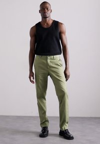 Groene katoen broek met een rechte pasvorm, gecombineerd met een zwart mouwloos topje. Het model draagt zwarte leren schoenen.