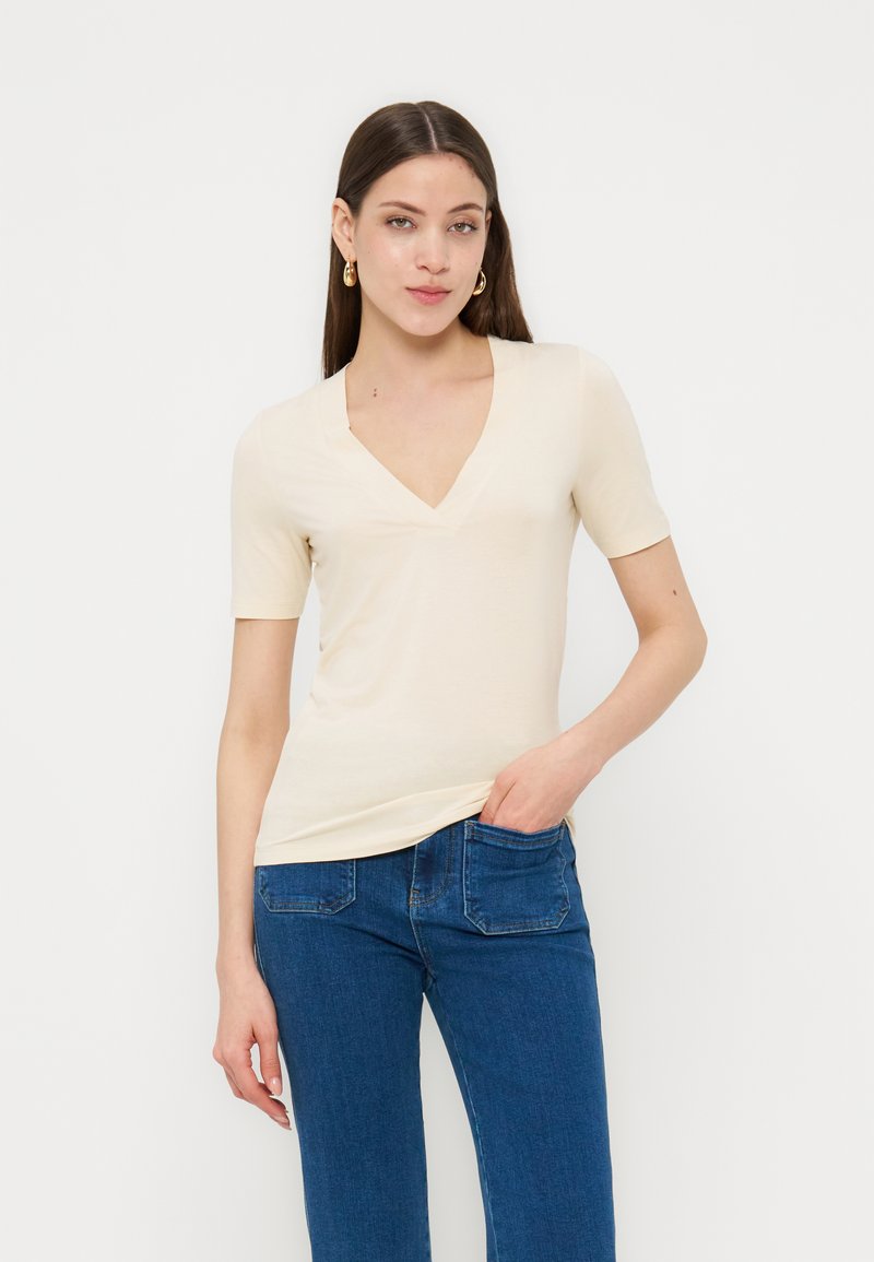 Νεαρή γυναίκα με μακριά καστανά μαλλιά, φορόντας χρυσούς κρίκους στα αυτιά, κρεμ μπλούζα με V λαιμόκοψη και κοντά μανίκια, και μπλε τζιν με το ένα χέρι στην τσέπη.