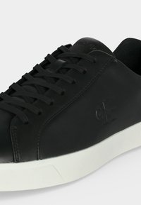 Μαύρο sneaker χαμηλής κοπής με μαύρα κορδόνια, λείο δερμάτινο πάνω μέρος, ανάγλυφο λογότυπο Calvin Klein στο πλάι και λευκή καουτσούκ σόλα.