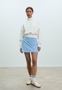 Biela mikina s dlhými rukávmi a vysokým golierom v kombinácii s svetlomodrou mini sukňou. Modelka má na sebe biele tenisky s čiernymi detailmi a ponožky.