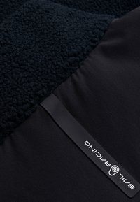 Tissu molleton noir avec une surface texturée et duveteuse, combiné à un matériau lisse et léger. Présente une étiquette de marque imprimée.