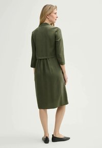 Olivengrünes Kleid mit Dreiviertelärmeln, Taillierung und knielangem Saum. Kombiniert mit schwarzen flachen Schuhen. Glatte Stoffoberfläche.