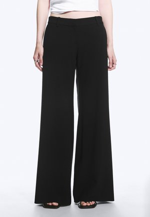 Personne portant un pantalon noir taille haute à jambes larges avec passants pour ceinture et sandales noires à bouts ouverts, debout sur fond blanc.