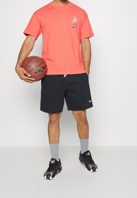 Coral short-sleeve T-shirt with a nautical design, paired with black shorts, gray socks, and black athletic shoes, holding a basketball.
