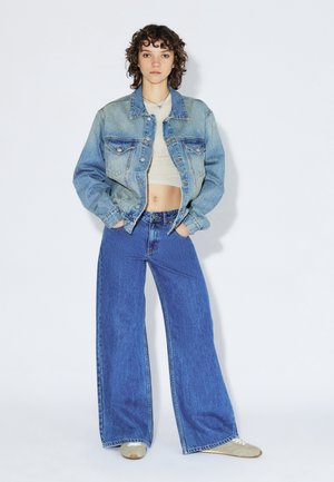 Denim jacket in light blue, cropped design with two chest pockets. Wide-leg blue jeans, high-waisted, with a smooth texture, paired with light sneakers.
