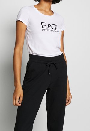 Vrouw draagt een wit EA7 Emporio Armani logot-shirt en zwarte broek met koordje tegen een effen lichte achtergrond.