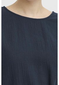 Närbild av en person som bär en mörk marinblå topp av texturerat tyg med rund halsringning, som visar nacken och övre bröstet.