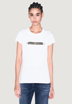 Giovane donna con i capelli intrecciati che indossa una maglietta bianca con testo decorato "ARMANI EXCHANGE" e jeans blu, in piedi davanti a uno sfondo semplice.