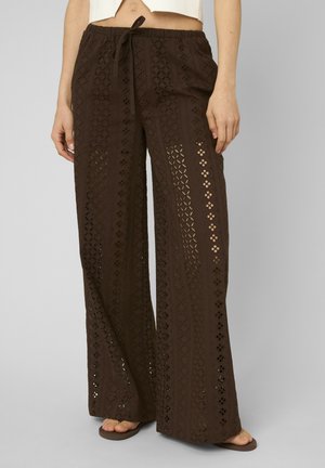 Pantalon large en crochet marron avec taille à cordon porté avec des tongs marron, montrant partiellement le ventre et les mains détendues sur les côtés.