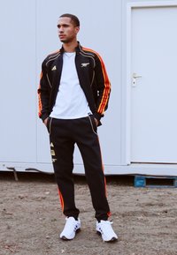 Black athletic tracksuit with red and yellow stripes, white t-shirt, and white sneakers. Pants feature zippered pockets and branding detail.