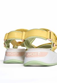 Sandalias amarillas y verdes con correas ajustables, detalles de suela blanca y un logo rosa en el talón. Texturas de cuero y tela suaves.