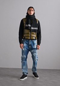 Puffer vest in shiny olive green, black sweater, distressed blue jeans with paint splatters, and black sneakers. Black scarf with logo.