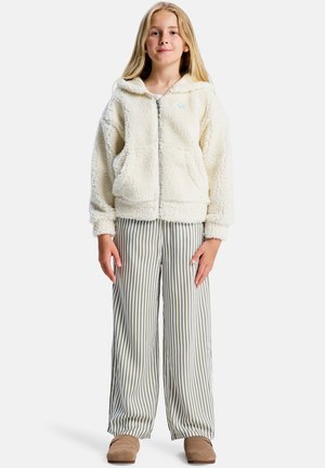 Beigefarbenes Fleece-Kapuzenjacke mit Reißverschluss und vorderen Taschen, kombiniert mit weichen, gestreiften, weiten Hosen in Grün und Weiß sowie braunen Slip-on-Schuhen.