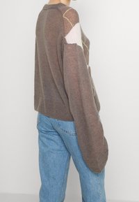Brun strikket sweater med en løs pasform, der har kontrasterende pastel geometriske mønstre og ribbede manchetter, parret med høj talje blå jeans.