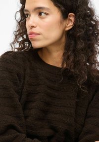 Donna con capelli ricci e scuri, che indossa un maglione scuro e strutturato, guarda verso sinistra con un'espressione neutra su uno sfondo semplice.