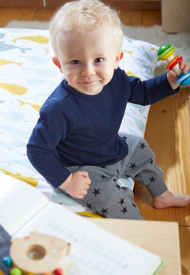 Child sitting on a patterned blanket, wearing a navy long-sleeve shirt and gray pants with black stars, holding a colorful rattle.