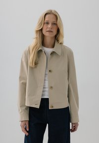 Blondynka w beżowej zapinanej kurtce na białej bluzce i ciemnych spodniach, stojąca na jednolitym jasnym tle.