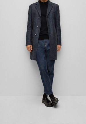 Cappotto texturizzato blu scuro con sei bottoni, abbinato a un dolcevita nero, jeans blu e scarpe nere. Design minimalista e vestibilità su misura.