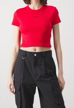 Kobieta w czerwonym krótkim T-shirtcie z logo "Hugo" oraz czarnych, wysokich spodni typu cargo z bocznymi kieszeniami i metalowym pierścieniem przy talii.