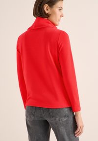 Rotes Langarmoberteil mit hohem Kragen, aus glattem Stoff gefertigt. Kombiniert mit grauen Jeans, die einen entspannten Schnitt und ein schlichtes Design aufweisen.