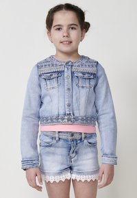 Svetlo modra denim jakna s skrajšano dolžino in dekorativnim šivanjem na ramenskem delu, z dvema sprednjima žepoma in srebrnimi gumbi. Usklajena s svetlo modrimi kratkimi hlačami.