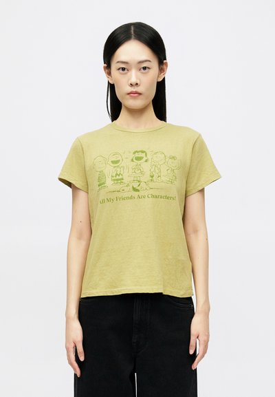 Giovane donna con capelli neri lisci, che indossa una maglietta verde con personaggi dei Peanuts e del testo, abbinata a pantaloni neri su uno sfondo bianco.