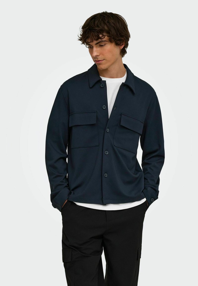 Jeune homme portant une veste-chemise bleu marine foncé boutonnée sur un t-shirt blanc et un pantalon cargo noir, debout les mains dans les poches.