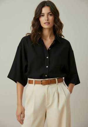 Vrouw met golvend bruin haar die een losse zwarte blouse met knopen draagt, beige broek met hoge taille en een bruine riem, staand met één hand in de zak.