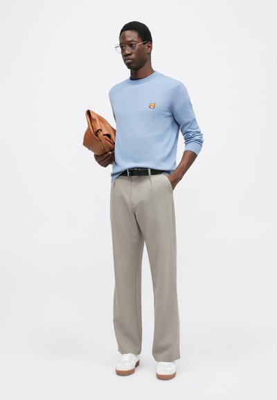 Pulover tricotat de culoare bleu deschis cu o aplicație în formă de vulpe, pantaloni bej cu croi tailored și adidași albi cu talpă din cauciuc, ținând o geantă din piele maro.