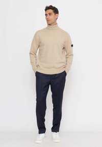Maglione beige a collo alto con polsini a coste, abbinato a pantaloni blu scuro e sneaker bianche; tessuto dalla texture liscia; design semplice.