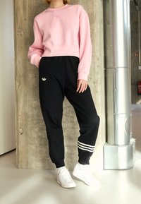 Osoba v růžovém svetru a černých teplákových kalhotách Adidas s bílými pruhy a bílých teniskách, stojící uvnitř vedle betonové zdi a kovového potrubí.