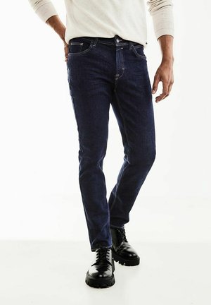 Man draagt donkerblauwe slim-fit jeans, zwarte veterlaarzen en een lichtbeige overhemd met lange mouwen, staand tegen een witte achtergrond.