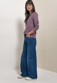 Femme debout, les mains dans les poches, portant un sweat à capuche violet à manches longues, un jean bleu à jambes larges et des baskets blanches sur un fond neutre.