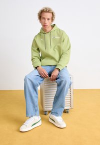 Sweatshirt à capuche vert avec poche frontale, jean bleu clair et baskets blanches avec accents verts, assis sur une boîte de rangement métallique.