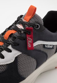 Nahaufnahme eines schwarzen, weißen und grauen Sneakers mit orangen Zuglaschen, schwarzen Schnürsenkeln, rotem Markenetikett und "s.Oliver"-Logo an der Seitenwand.