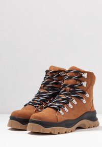 Bottes de randonnée montantes en daim marron avec des lacets rayés noirs et blancs, des œillets métalliques et une semelle en caoutchouc robuste. Tige texturée pour plus de durabilité.