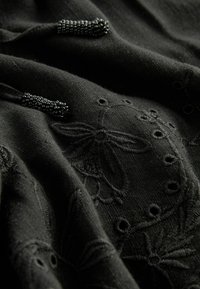 Tissu noir avec broderie florale en relief et découpes circulaires ; présente des extrémités de lien décoratives en perles pour plus de détail et de texture.