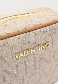 Kosmetyczka Valentino wykonana z beżowego materiału o fakturze, z złotym zamkiem i logo. Posiada gładki jasnobrązowy akcent wzdłuż szwów.