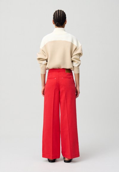Suéter oversized en bloques de color beige y blanco con un alto cuello, combinado con pantalones anchos de color rojo brillante y zapatos negros.