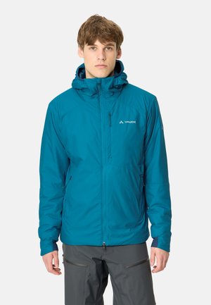 Jonge man die een blauwe Vaude jas met capuchon en donkergrijze broek draagt, staand tegen een effen witte achtergrond.