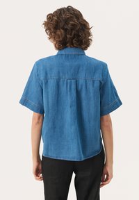 Camicia di jeans a maniche corte, azzurro chiaro, con cucitura sul retro e vestibilità ampia. I bordi presentano cuciture in contrasto. Indossata con pantaloni neri.