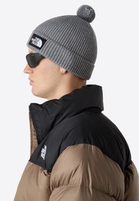 Šedá pletená čepice s pomponem a černou značkovou nášivkou, v kombinaci s černou a hnědou prošívanou bundou a tmavými slunečními brýlemi.