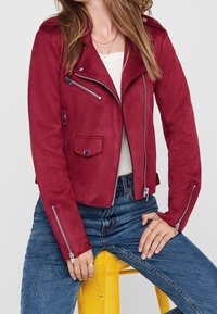 Femme portant une veste en suède rouge zippée, un haut blanc, un jean bleu et des colliers en or, assise sur un tabouret en métal perforé jaune.