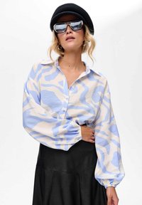 Vrouw in een oversized blouse met blauwe en witte patronen, zwarte rok, zwarte baret, grote zonnebril en ringoorbellen tegen een witte achtergrond.