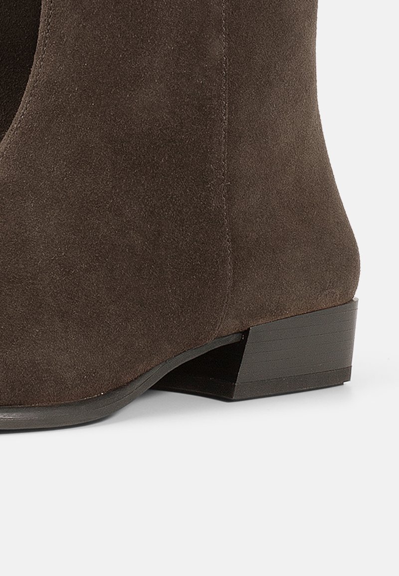 Bottine en suede marron avec un petit talon carré, un bout arrondi et un détail de découpe sur le côté pour un design supplémentaire. Texture lisse sur l'ensemble.