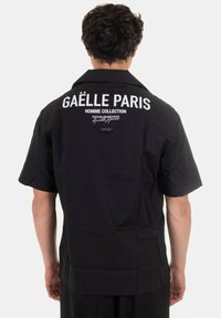 Maglietta nera a maniche corte con un logo bianco prominente sulla schiena, con una trama liscia e colletto a camicia.