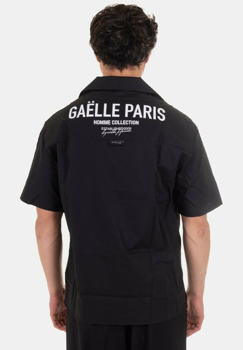 Maglietta nera a maniche corte con un logo bianco prominente sulla schiena, con una trama liscia e colletto a camicia.