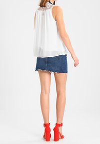 Witte mouwloze blouse met een gestructureerde kant achterzijde, gecombineerd met een denimrok met rafelige zoom en helderrode blokhakken.