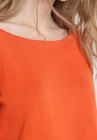 Haut en tricot léger orange, avec un large col et une texture côtelée, mettant en valeur un design ajusté qui accentue les épaules.