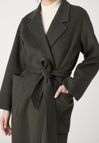 Abrigo de lana verde oscuro con cinturón de lazo, solapas de muesca y bolsillo de parche. Cuenta con mangas largas y una textura suave.